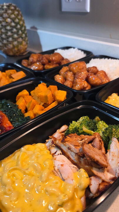 Meal prep organized in black containers: grilled chicken, creamy mac and cheese, bright orange squash, sautéed greens, sweet orange chicken, and white rice, with a pineapple visible in the background.